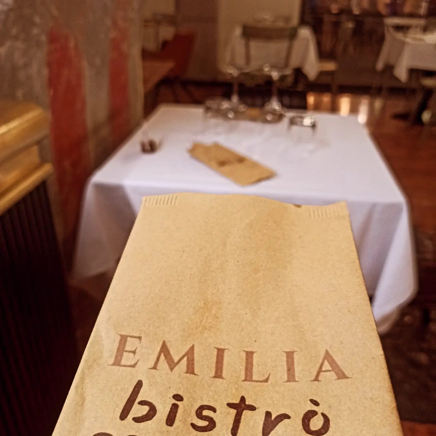 Tavolo apparecchiato con tovaglia bianca e il copriposate in carta kraft con il marchio Emilia bistrò, nella sala di Emilia Bistrò, Modena.