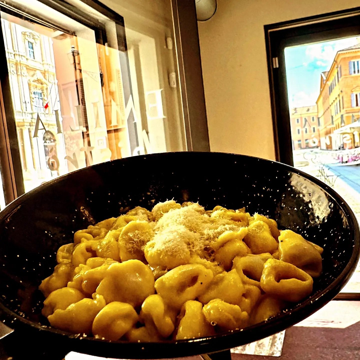 Ciotola di tortellini con Parmigiano grattugiato, sul davanzale: dietro, il Palazzo Ducale visto attraverso la vetrina di Emilia Bistrò.